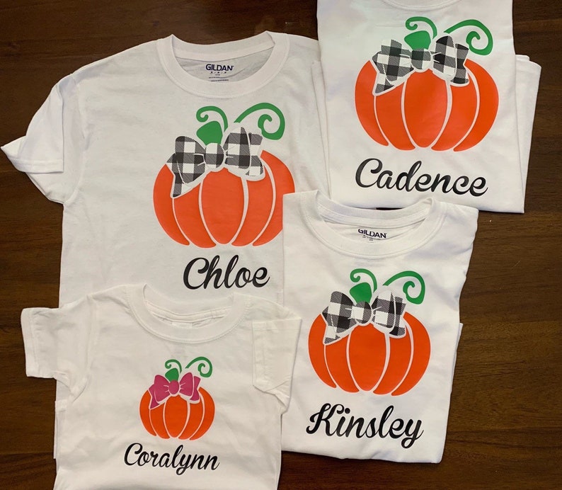 May include: Four white t-shirts with a pumpkin design and a name on each. The pumpkins have a black and white checkered bow. The names are Coralyann, Chloe, Kinsley, and Cadence.