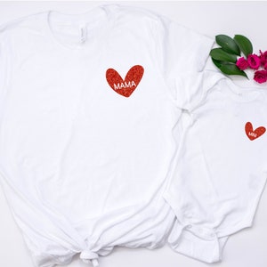 May include: Two white t-shirts with red glitter hearts. The larger shirt has the word "MAMA" inside the heart. The smaller shirt has a smaller heart with no text.