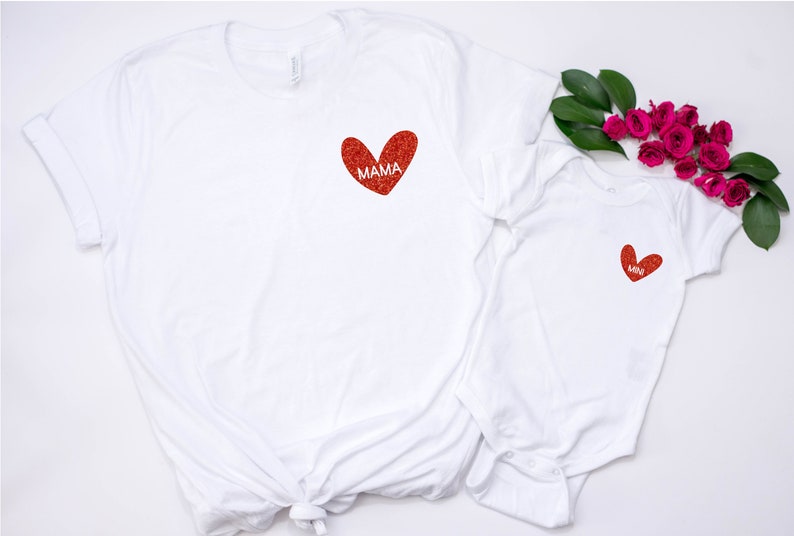 May include: Two white t-shirts with red glitter hearts. The larger shirt has the word "MAMA" inside the heart. The smaller shirt has a smaller heart with no text.
