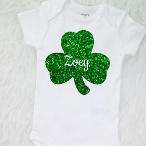 May include: White baby onesie with a green glitter shamrock and the name "Zoey" in white.