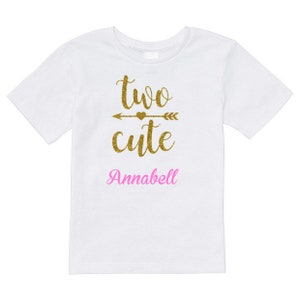 May include: White t-shirt with gold glitter text that says "two cute" with an arrow and heart above the word "cute". The name "Annabell" is written in pink glitter below the text.