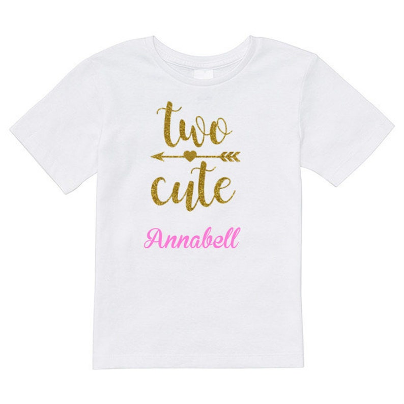 May include: White t-shirt with gold glitter text that says "two cute" with an arrow and heart above the word "cute". The name "Annabell" is written in pink glitter below the text.
