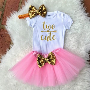 May include: A pink tulle tutu with a gold sequin bow, a white onesie with the text "two cute" in gold glitter, and a gold sequin bow headband.