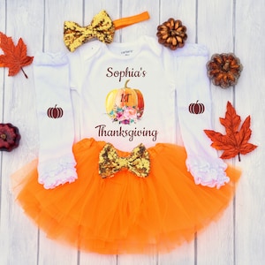 May include: Orange tulle skirt with a gold sequin bow, a white long-sleeved onesie with a pumpkin graphic and the text "Sophia's 1st Thanksgiving", and a gold sequin headband.