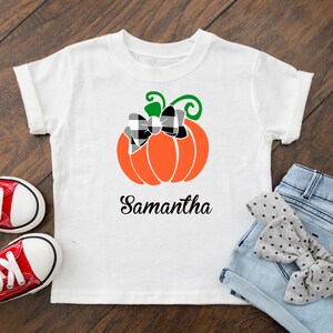 May include: White t-shirt with a black and white checkered bow on an orange pumpkin with a green stem. The text "Samantha" is printed below the pumpkin.