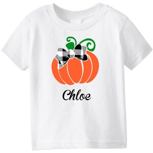 May include: White t-shirt with a black and white checkered bow on an orange pumpkin. The text "Chloe" is printed below the pumpkin.