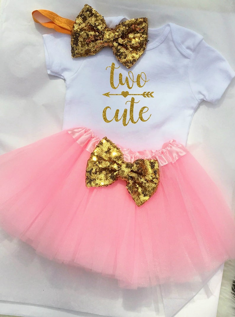 May include: A pink tulle tutu with a gold sequin bow, a white onesie with gold glitter text that says "two cute" and a gold sequin bow headband.