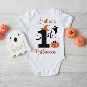 May include: White baby onesie with orange glitter text that says "Sophia's 1st Halloween" with a black number one, a ghost, a bat, and a pumpkin.