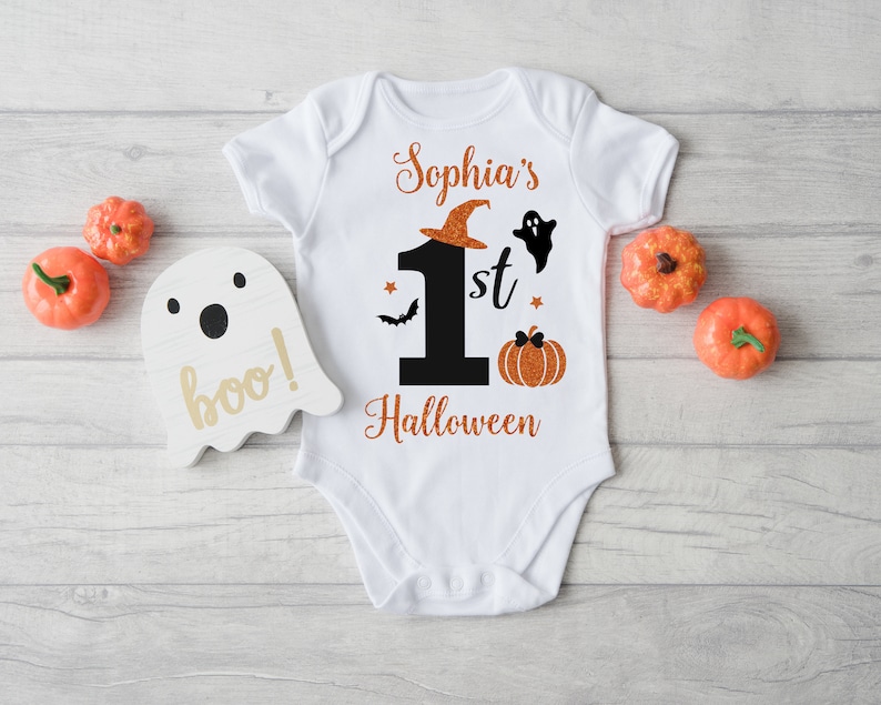 May include: White baby onesie with orange glitter text that says "Sophia's 1st Halloween" with a black number one, a ghost, a bat, and a pumpkin.