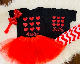 May include: Two black baby onesies with red glitter hearts and the names "Kenley" and "Bella" printed on them. One onesie is paired with a red tulle tutu and a red glitter bow headband. The other onesie is paired with red and white chevron leggings.