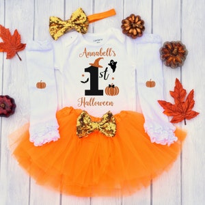 May include: Orange and white Halloween baby outfit with a white long-sleeved onesie, orange tulle skirt, and a gold glitter bow. The onesie has the text "Annabell's 1st Halloween" with a black number one, a witch's hat, a ghost, a bat, and a pumpkin.