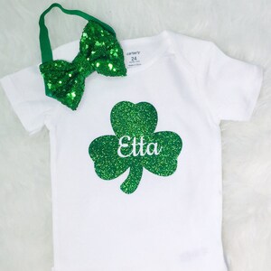 May include: White bodysuit with a green glitter shamrock and the name "Etta" on it. A green glitter bow headband is included.