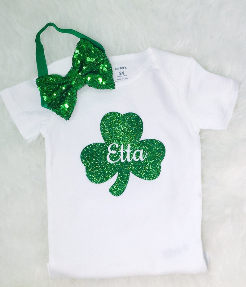 May include: White bodysuit with a green glitter shamrock and the name "Etta" on it. A green glitter bow headband is included.