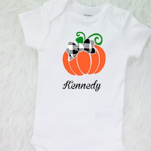 May include: White onesie with a black and white checkered bow on an orange pumpkin with a green stem. The onesie has the name "Kennedy" printed below the pumpkin.