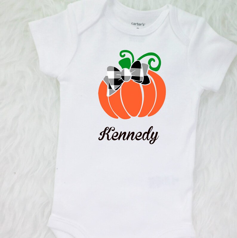 May include: White onesie with a black and white checkered bow on an orange pumpkin with a green stem. The onesie has the name "Kennedy" printed below the pumpkin.