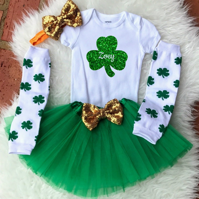 May include: A green and gold St. Patrick's Day outfit for a baby girl. The outfit includes a white onesie with a green shamrock and the name "Zoey" on it, a green tulle skirt with a gold bow, white leg warmers with green shamrocks, and a gold headband with a bow.