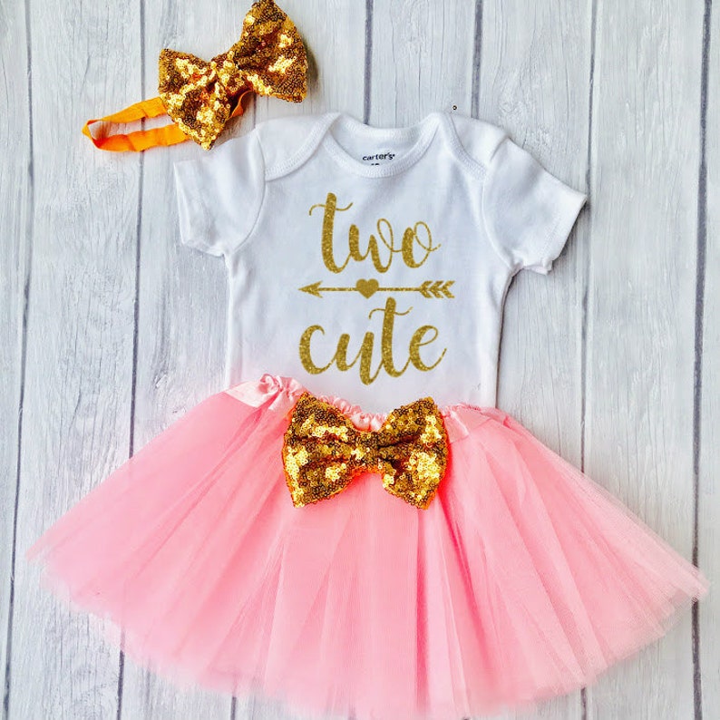 May include: A pink tulle tutu skirt with a gold sequin bow, a white onesie with the text "two cute" in gold glitter, and a gold sequin bow headband.