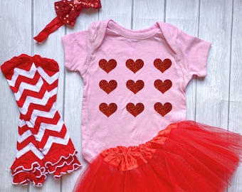 May include: A baby girl's Valentine's Day outfit set. The set includes a pink onesie with nine red glitter hearts, a red tutu skirt, red and white chevron leg warmers, and a red sequin bow headband.