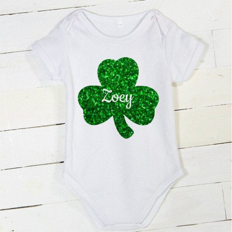 May include: White baby onesie with a green glitter shamrock and the name "Zoey" printed on it.