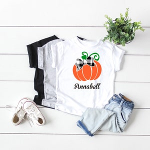 May include: White t-shirt with a black and white checkered bow on an orange pumpkin with green stem and leaves. The text "Annabell" is printed below the pumpkin.
