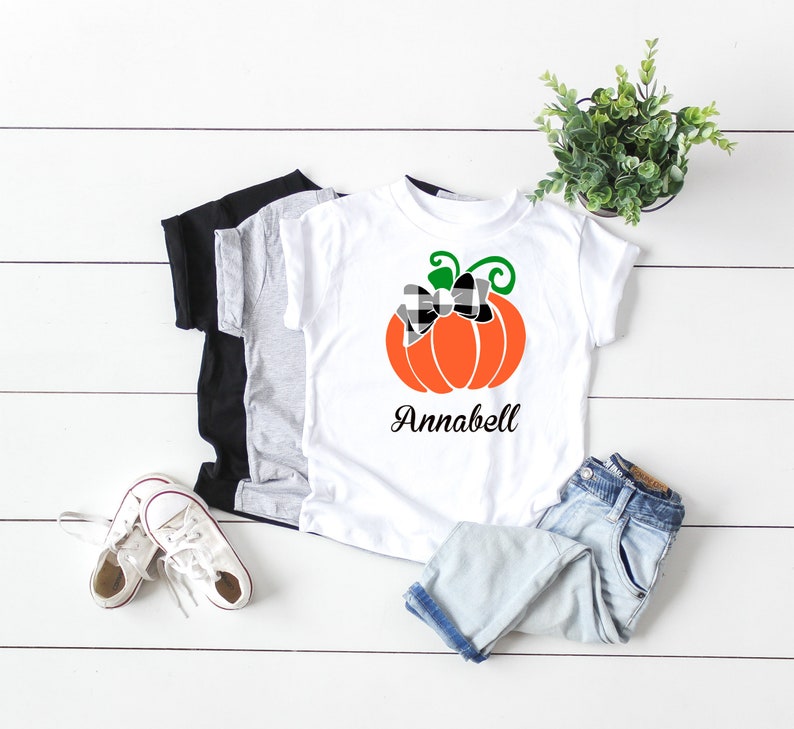 May include: White t-shirt with a black and white checkered bow on an orange pumpkin with green stem and leaves. The text "Annabell" is printed below the pumpkin.