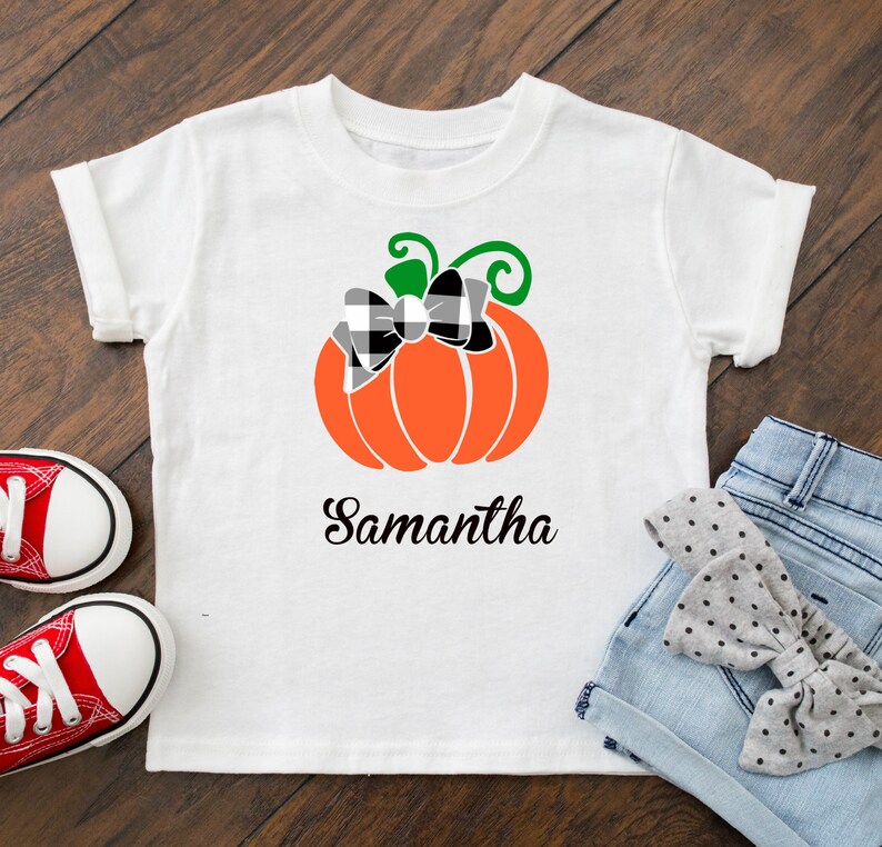 May include: White t-shirt with a black and white checkered bow on an orange pumpkin with a green stem. The text "Samantha" is printed below the pumpkin.