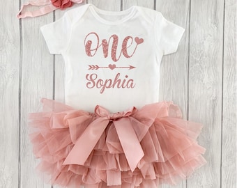 May include: A white short-sleeved bodysuit with rose gold glitter text reading One and the name Sophia, featuring an arrow and heart graphic. The set includes a dusty rose layered tulle tutu skirt with a satin bow and a matching floral headband.