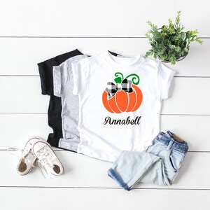 May include: White t-shirt with a black and white checkered bow on an orange pumpkin with green stem and leaves. The text "Annabell" is printed below the pumpkin.