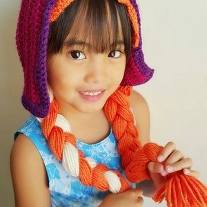 May include: A young girl wearing a purple, orange, and pink crocheted hat with a long orange and white braided yarn attached. The girl is wearing a blue dress with a snowflake pattern.