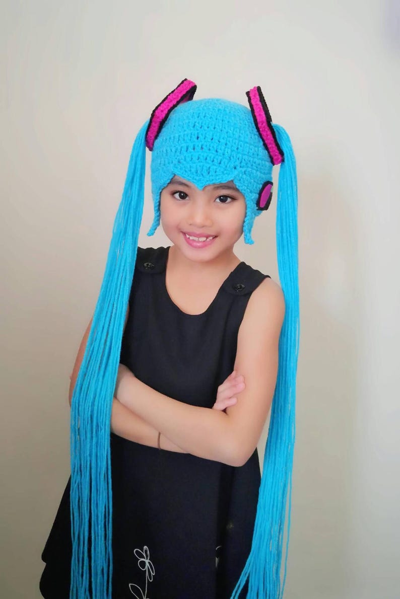 May include: A young person wearing a blue crocheted hat with long blue yarn tails, pink and black accents, and a black dress.