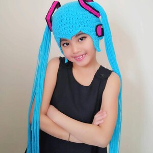 May include: A young person wearing a blue crocheted hat with long blue yarn tails. The hat has pink and black accents and resembles a popular anime character.