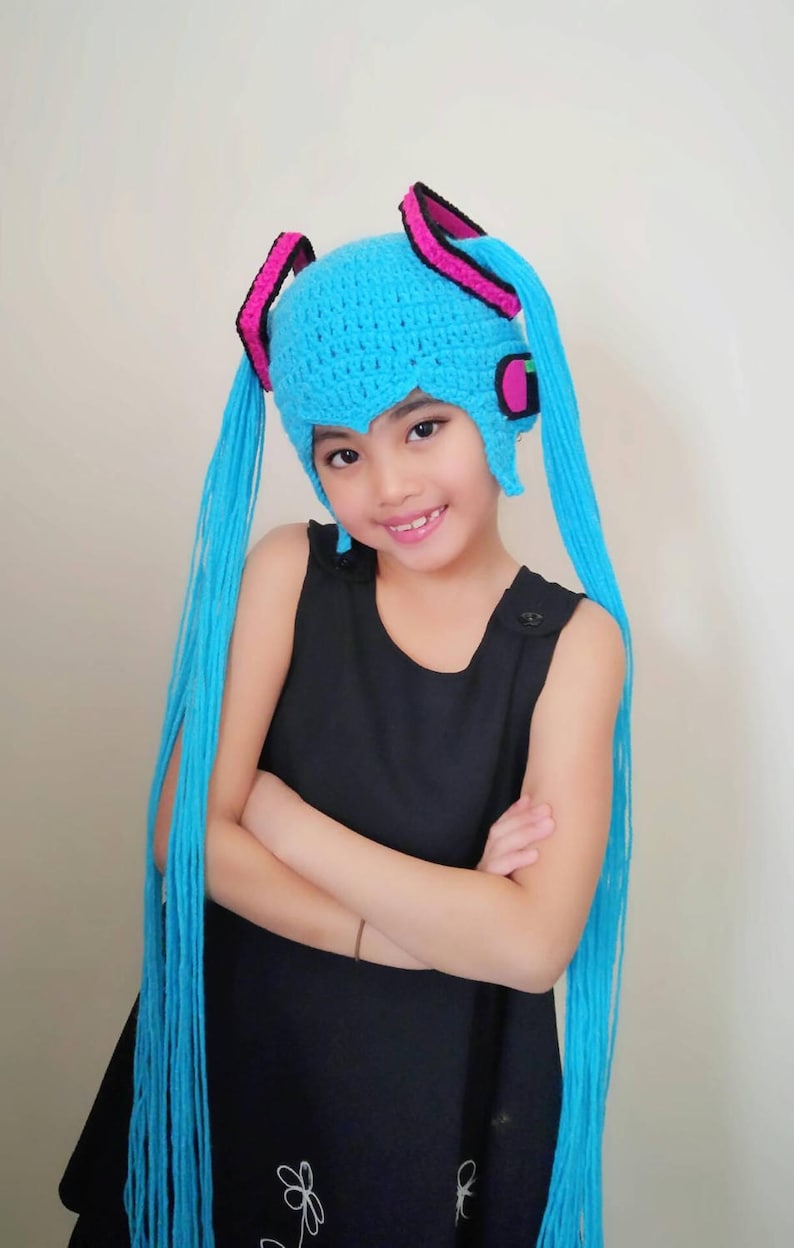 May include: A young person wearing a blue crocheted hat with long blue yarn tails. The hat has pink and black accents and resembles a popular anime character.