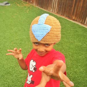 Puede incluir: Un niño lleva un gorro de punto marrón con un diseño de flecha azul. El gorro recuerda al personaje de Avatar: La Leyenda de Aang, Aang. El niño lleva una camiseta roja con un diseño de mano de dibujos animados blanco.