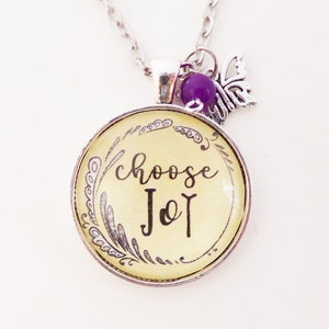 May include: A silver necklace with a round pendant that says "choose joy" in black script. The pendant is surrounded by a silver frame with a decorative design. A small purple bead and a silver butterfly charm hang from the necklace.