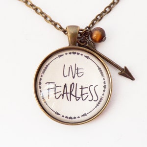 May include: A bronze necklace with a round pendant that says "Live Fearless" in black lettering. The pendant is surrounded by a border of small arrows and hearts. The necklace also has a small arrow charm and a brown bead.