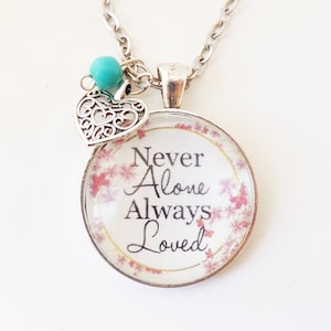 May include: Silver necklace with a round pendant featuring the text "Never Alone Always Loved" on a floral background. The pendant is attached to a silver chain with a small turquoise bead and a filigree heart charm.
