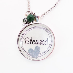 May include: Silver-toned necklace with a round pendant. The pendant features the word "Blessed" in cursive, a blue heart, and a decorative border. A green bead and floral accents adorn the top of the pendant.