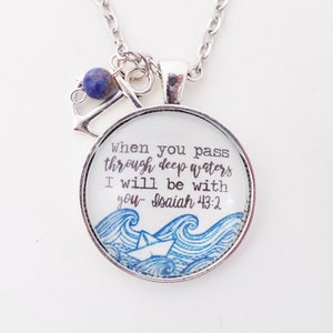 May include: Silver necklace with a round pendant featuring a blue and white illustration of waves and a paper boat. The pendant has the text "When you pass through deep waters I will be with you - Isaiah 43:2".
