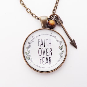May include: A bronze necklace with a round pendant featuring the words "Faith Over Fear" surrounded by a leafy wreath. A small arrow charm hangs from the pendant.