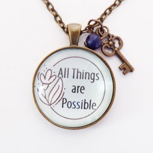 Puede incluir: Un collar de bronce con un colgante redondo de vidrio que dice "All Things are Possible". El colgante tiene un pequeño diseño de flor y está unido a una cadena de bronce con un pequeño dije de llave y una cuenta morada.