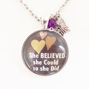 May include: A silver necklace with a round pendant featuring three gold hearts and the text "She BELIEVED she Could so she Did". The pendant has a purple gemstone charm and a silver butterfly charm.