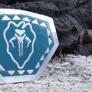 May include: A shield with a teal center and a silver border. The center features a white emblem of a stylized skull-like creature. The border is decorated with white triangles. The shield is resting on a textured surface.