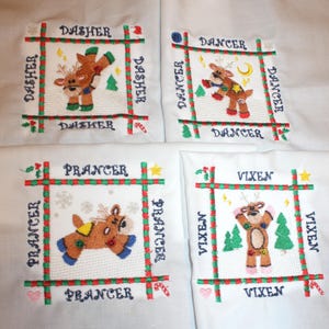May include: Four white fabric squares embroidered with reindeer designs and names. Each square features a different reindeer: Dasher, Dancer, Prancer, and Vixen. The reindeer are brown with colorful accents, surrounded by a red and green border with text.
