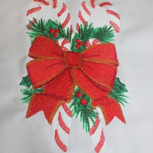 May include: Embroidered Christmas design featuring two candy canes with red and white stripes, green pine needles, and a large red bow with gold trim. The design is on a white background.