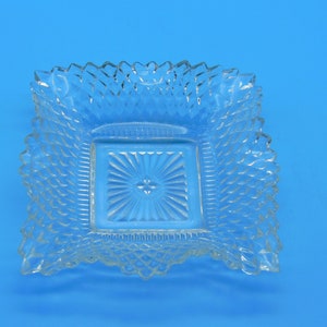 May include: Clear glass square dish with a textured, diamond-like pattern. The dish features a smaller, recessed square in the center with a sunburst design. The edges of the larger square have a scalloped, decorative trim. The overall style is vintage or antique, suggesting a decorative or serving piece.