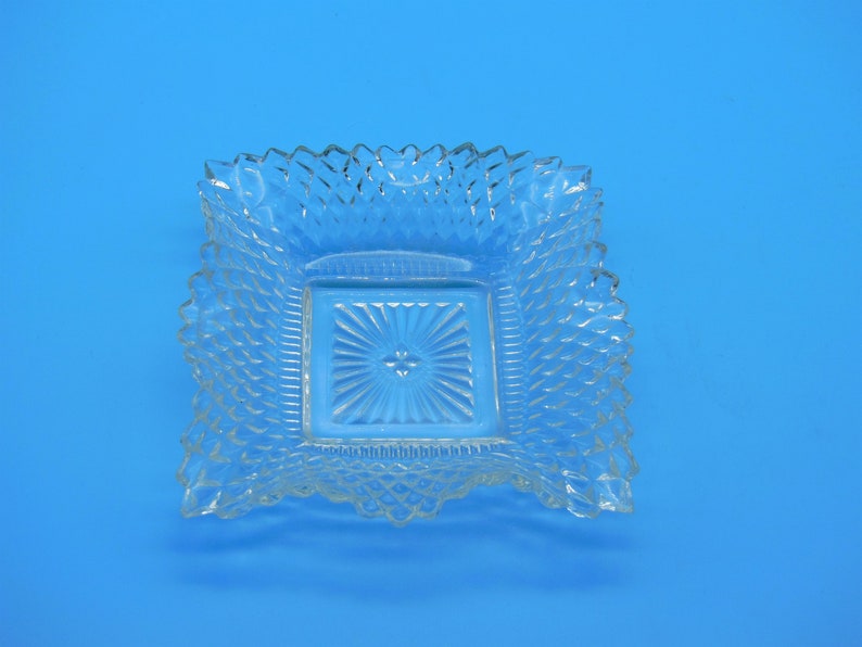 May include: Clear glass square dish with a textured, diamond-like pattern. The dish features a smaller, recessed square in the center with a sunburst design. The edges of the larger square have a scalloped, decorative trim. The overall style is vintage or antique, suggesting a decorative or serving piece.