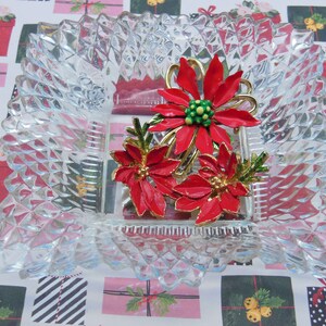 Puede incluir: Un broche de Navidad vintage con tres flores de poinsettia rojas con detalles dorados y hojas verdes. El broche se muestra en un plato de vidrio transparente.