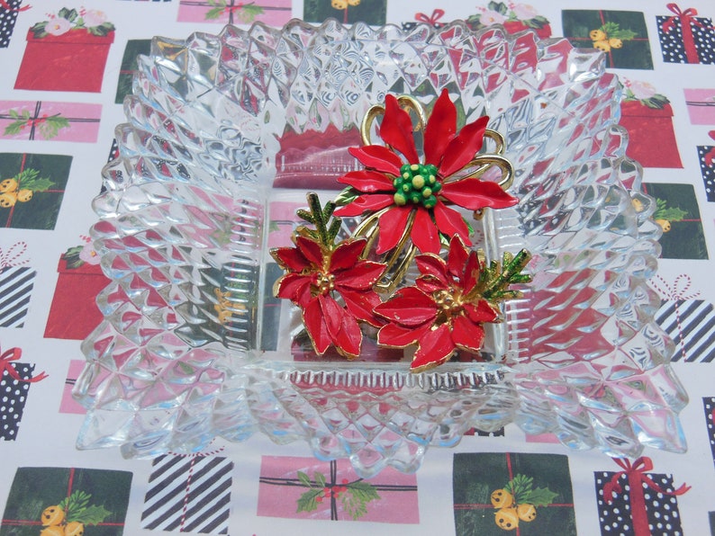 Puede incluir: Un broche de Navidad vintage con tres flores de poinsettia rojas con detalles dorados y hojas verdes. El broche se muestra en un plato de vidrio transparente.