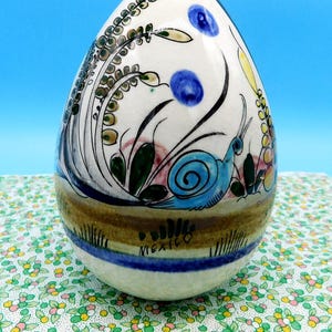 May include: A hand-painted ceramic egg with a floral and snail design. The egg features a white base with blue, green, and brown accents. The word "MEXICO" is written on the base.