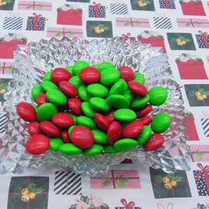 Puede incluir: Un taz&oacute;n de vidrio transparente lleno de caramelos M&M's rojos y verdes. El taz&oacute;n est&aacute; sobre un fondo blanco con un patr&oacute;n repetitivo de cajas de regalo rojas y verdes.